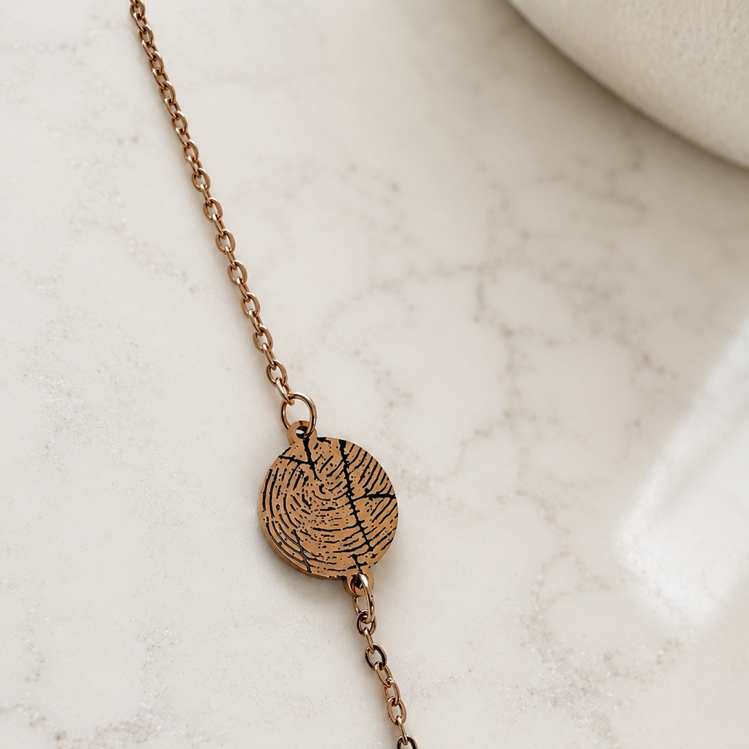 A delicate rose gold chain necklace featuring a small circular pendant engraved with a fingerprint pattern, resting on a light marble surface.