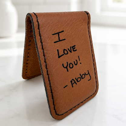 A brown leather wallet standing open on a light marble surface, with black stitched edges and handwritten text reading 'I Love You! - Abby' on the front.