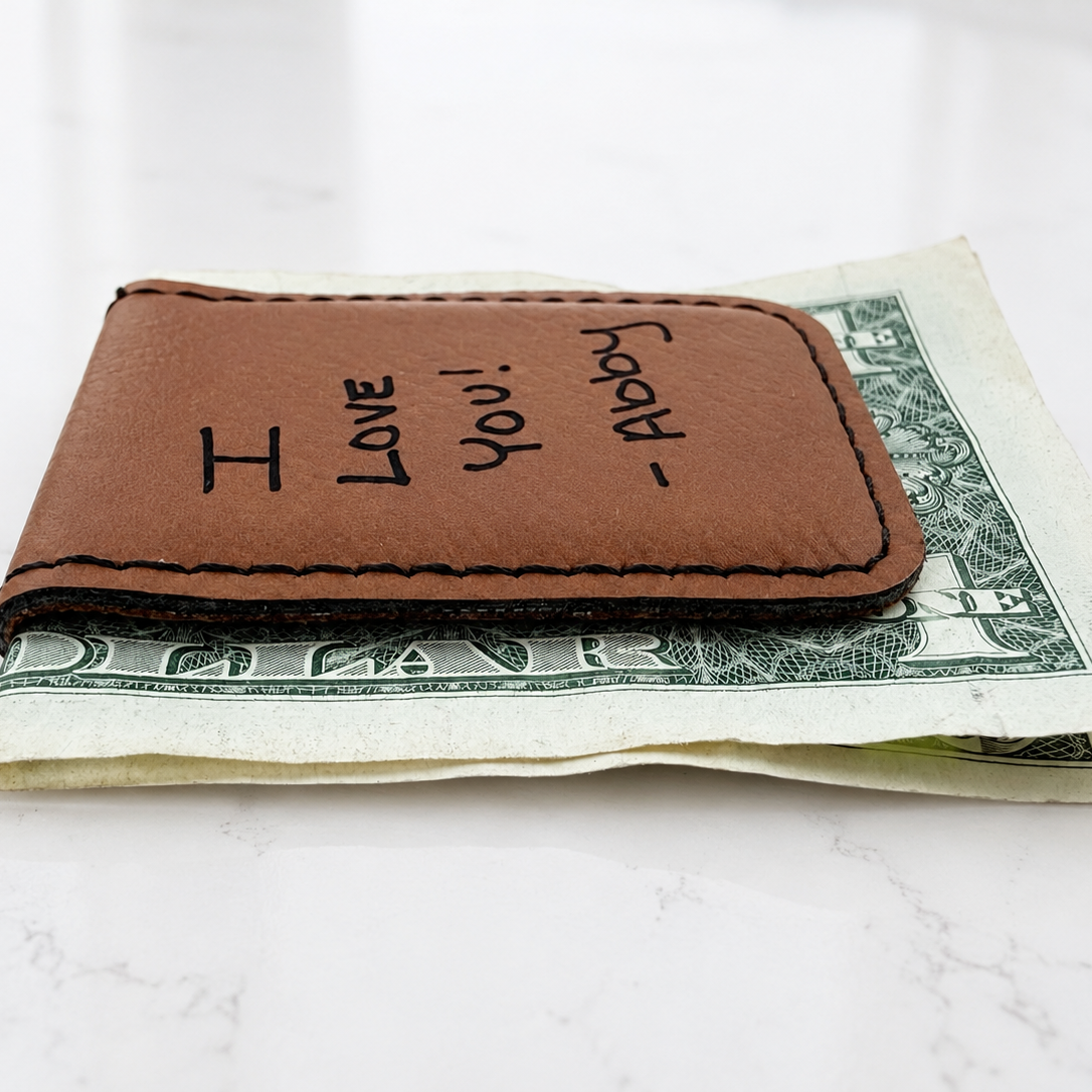 A brown leather money clip wallet with 'I LOVE You! ' Engraved. Holding a one-dollar bill on a white marble surface.