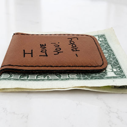 A brown leather money clip wallet with 'I LOVE You! ' Engraved. Holding a one-dollar bill on a white marble surface.