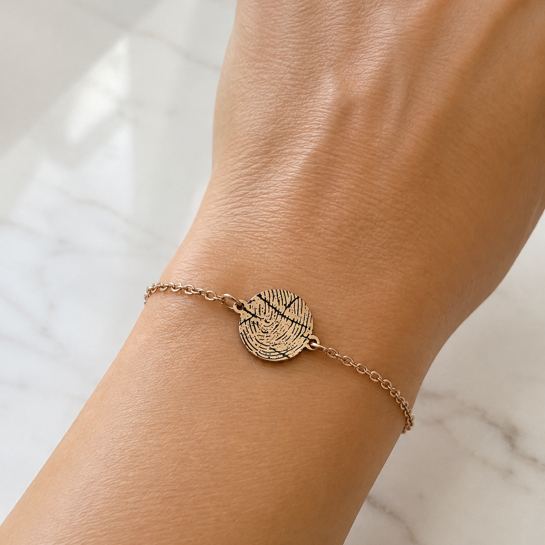 A delicate gold chain bracelet with a textured, engraved oval disc charm, shown worn on a person's wrist against a light marble background.