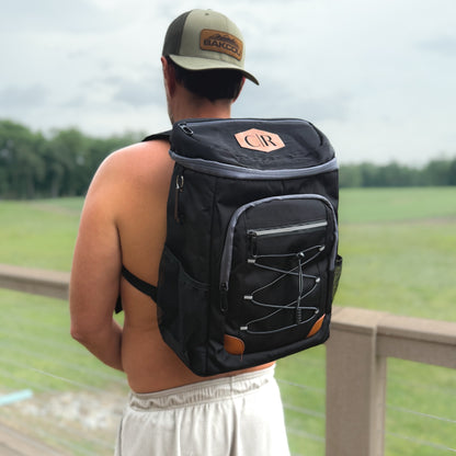 A man viewed from behind wearing a black backpack with gray trim and brown accents, standing outdoors on a wooden deck with a grassy field in the background. The backpack has a top zippered compartment, a front zippered pocket with elastic cord detail, and a small tan 'CR' patch on the top.
